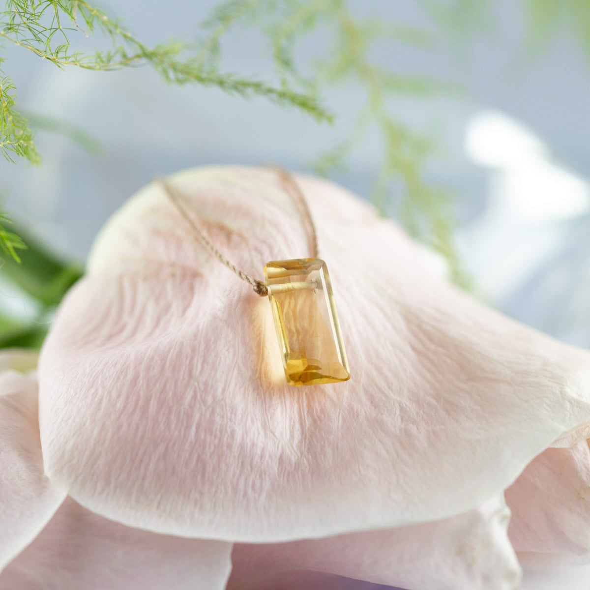 The Citrine Refined Necklace for Manifest by SoulKu shines atop a soft pink flower petal with green leaves and a blurred backdrop, perfect for handmade self-expression.