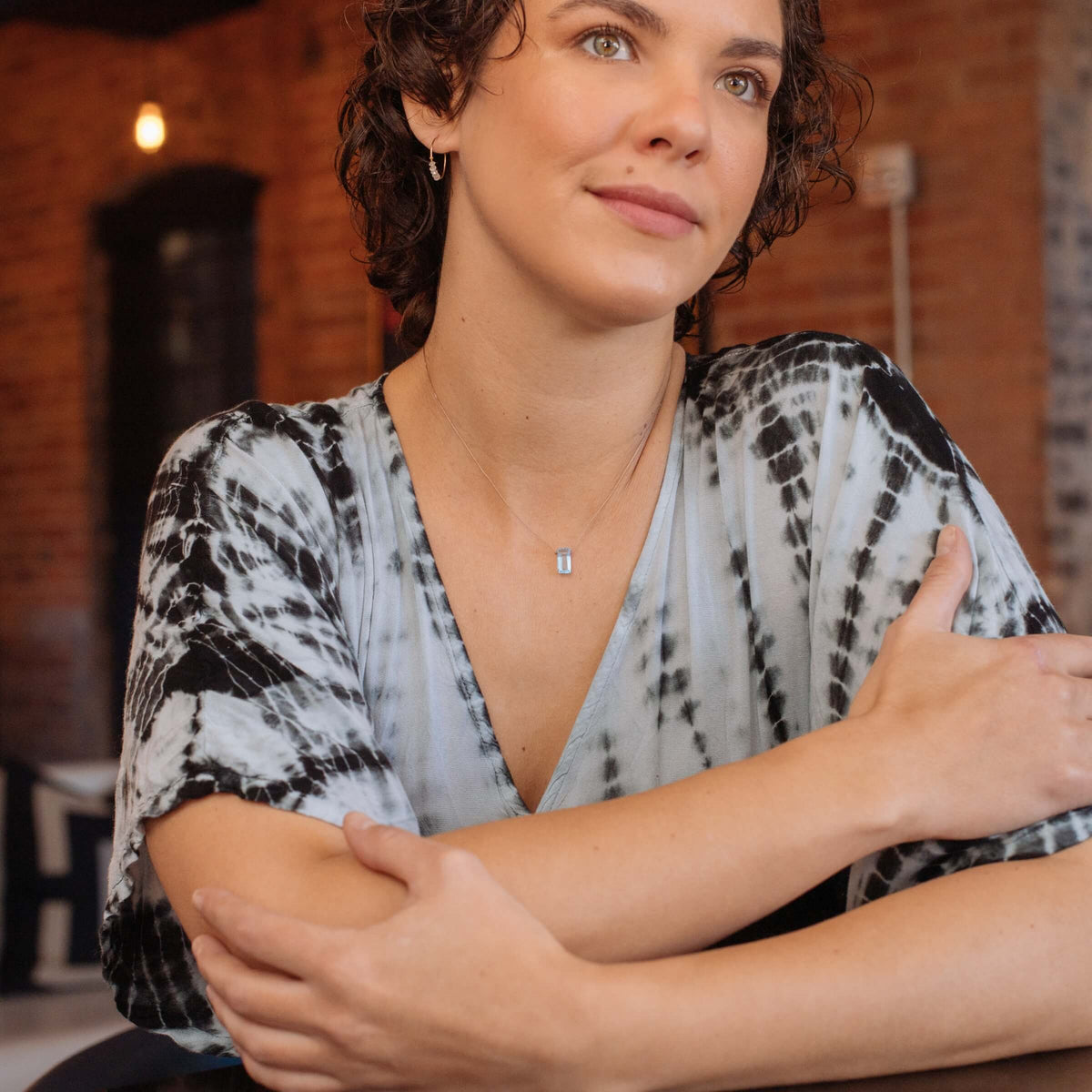A woman with short curly hair wears a tie-dye shirt and the SoulKu Sky Blue Topaz Refined Necklace for New Beginnings, sitting thoughtfully with arms crossed. A brick wall and warm light form the background.