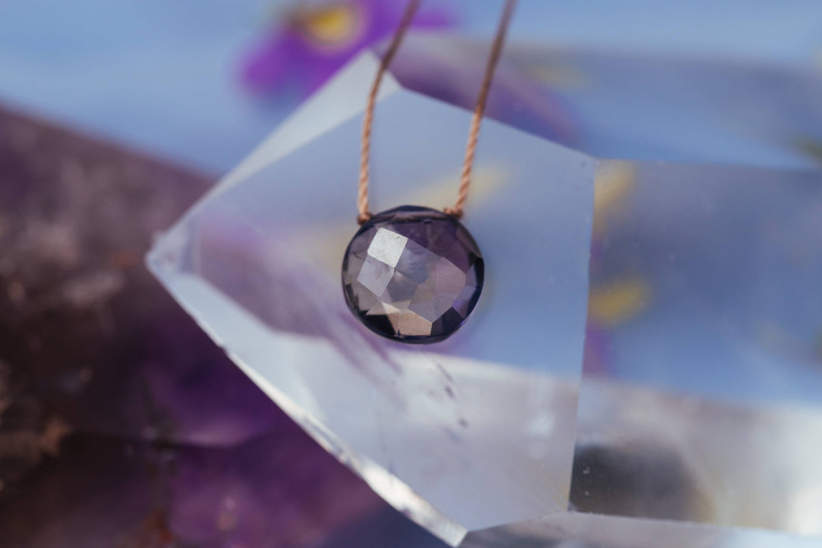 The Iolite Beam Necklace for ‘I Believe in You’ by SoulKu, featuring a faceted dark iolite gemstone on a tan cord, rests on clear quartz—radiating clarity and intuition with purple-blue hues in the background.