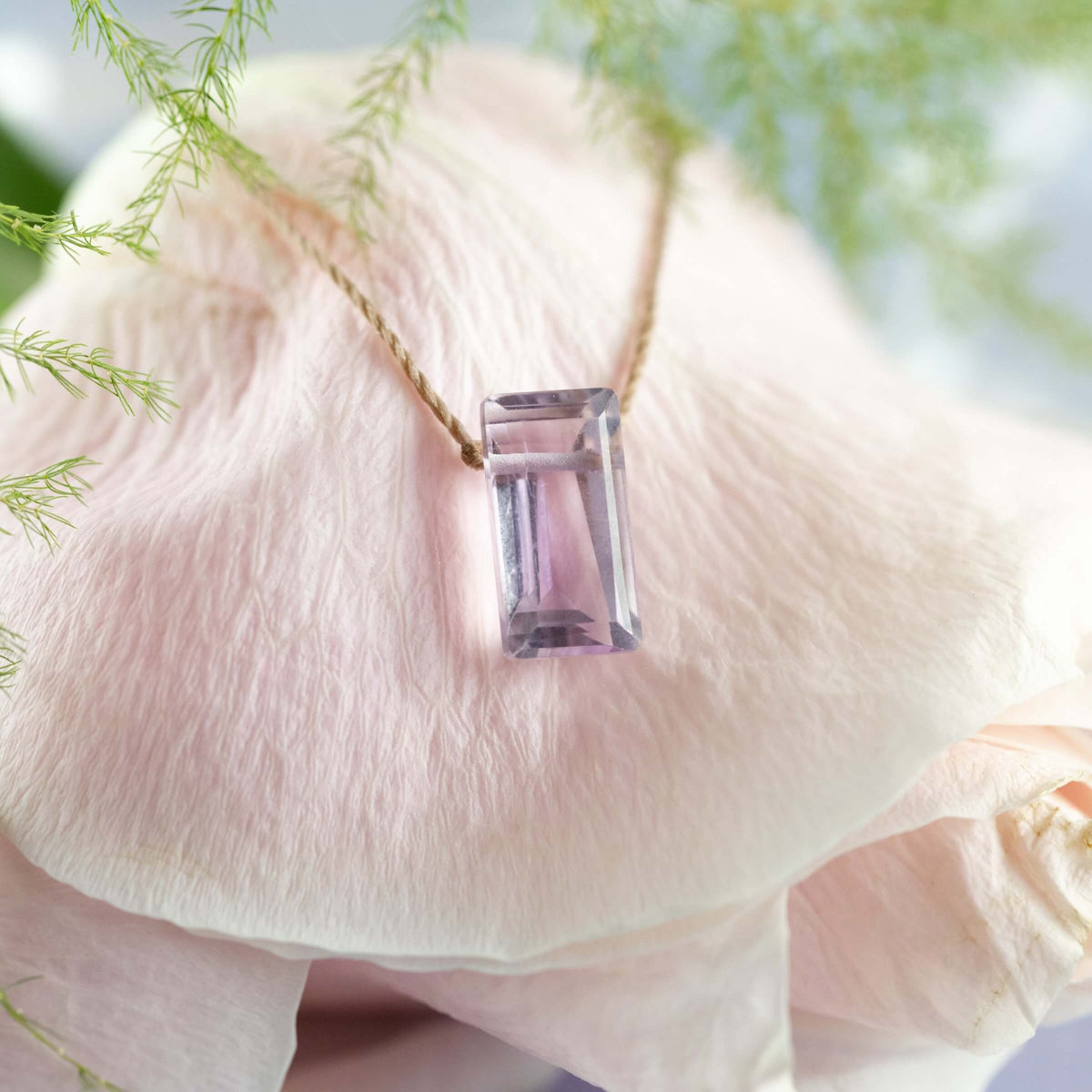 The Amethyst Refined Necklace for Fearless by SoulKu, featuring a rectangular faceted gemstone pendant on a chain, rests on a pale pink flower petal amid delicate green foliage.
