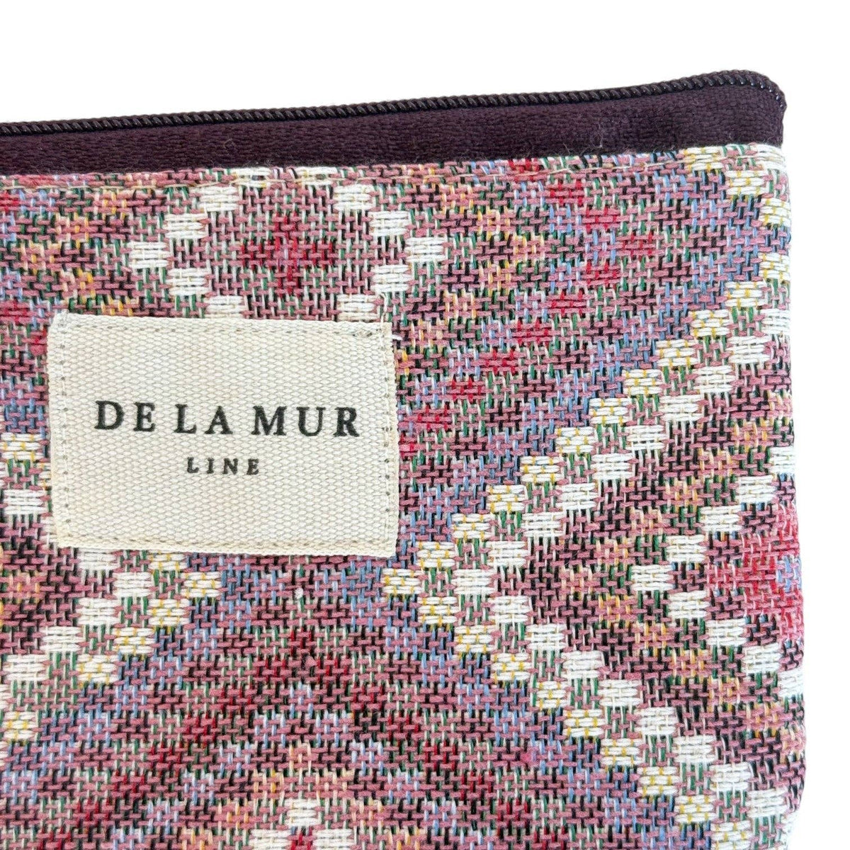 A detailed view of the Geometric Jacquard Coin Purse – Pink & Lilac (Nana) by DE LA MUR LINE US Inc highlights its intricate kilim-inspired pattern in pink, lilac, white, and yellow jacquard fabric. The front of the pouch is adorned with a beige DE LA MUR LINE label sewn on as a finishing touch.