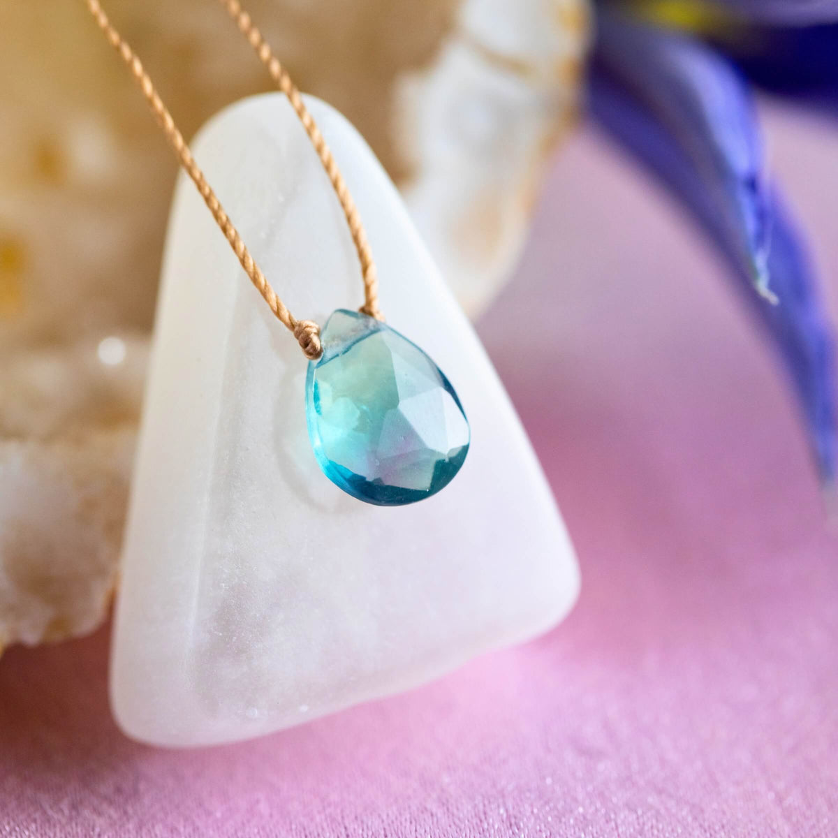 The Fluorite Luxe Necklace for Magic by SoulKu features a teardrop-shaped blue gemstone pendant on a light brown cord, displayed on a white stone with blurred flowers—unique handmade gemstone jewelry.