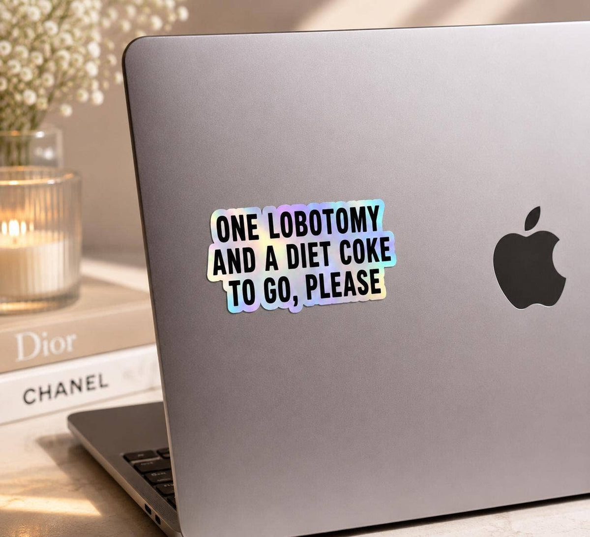 A laptop displays the Funny Diet Coke Sticker - Lobotomy to go from Hello Brand | Sticker My Art, with books and a candle softly blurred in the background.