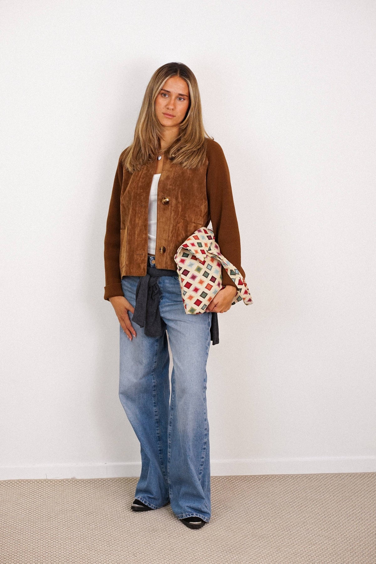 A woman with straight blonde hair and a neutral expression stands against a white wall, dressed in a brown jacket, white top, flared blue jeans, and black shoes. She is holding the DE LA MUR LINE US Inc Geometric Jacquard Medium Zippered Tote Bag – Multicolor (Zac).