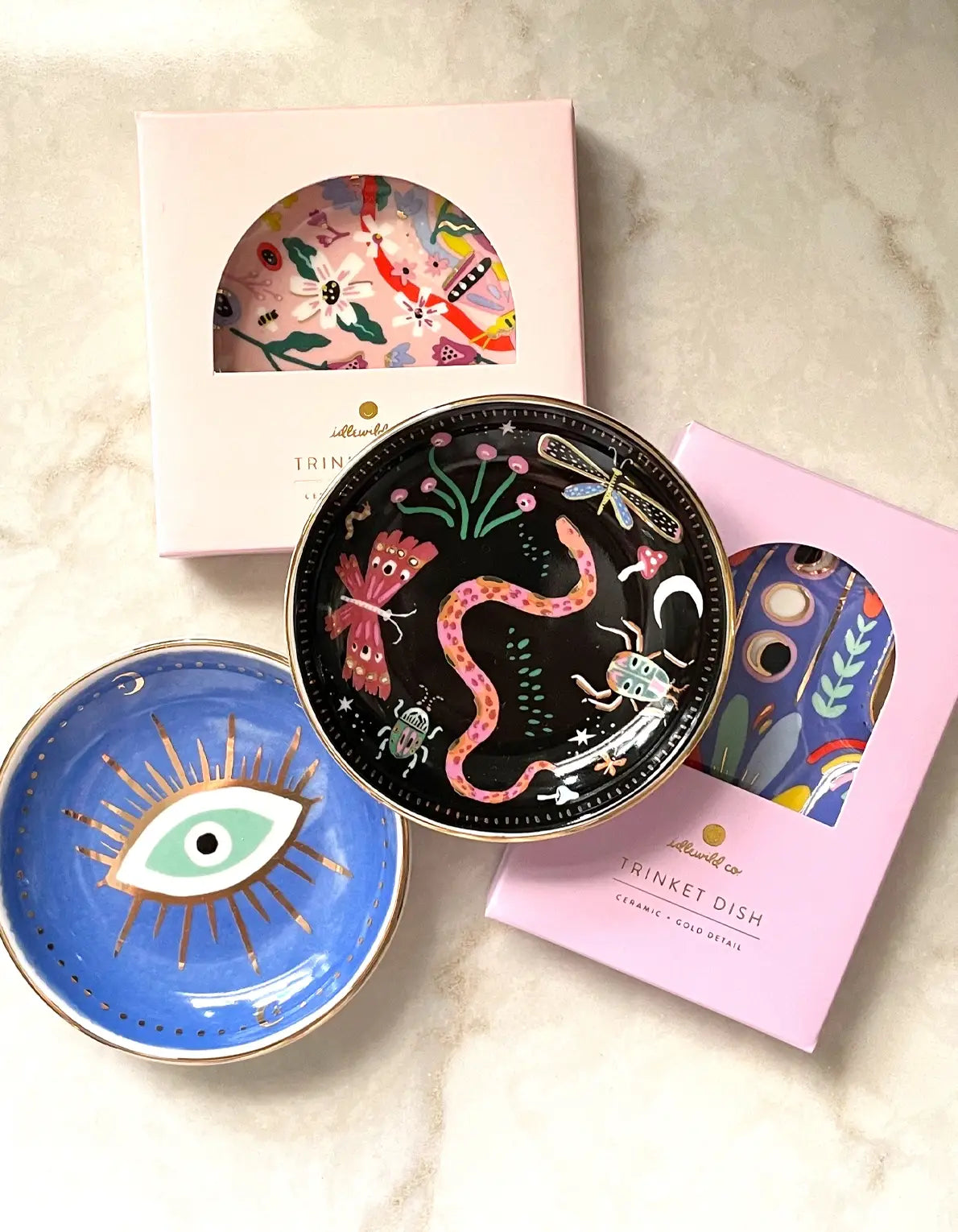 Two gold-edged Porcelain Trinket Dish - Evil Eye ceramic catchall dishes: one blue with an evil eye design and one black featuring snake and insect illustrations, displayed with their respective pink and white windowed gift boxes.