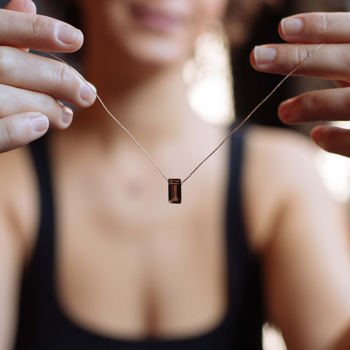A person holds the SoulKu Smoky Quartz Refined Necklace for Release, featuring a small cylindrical pendant. The person, wearing a black top, is out of focus in the background.