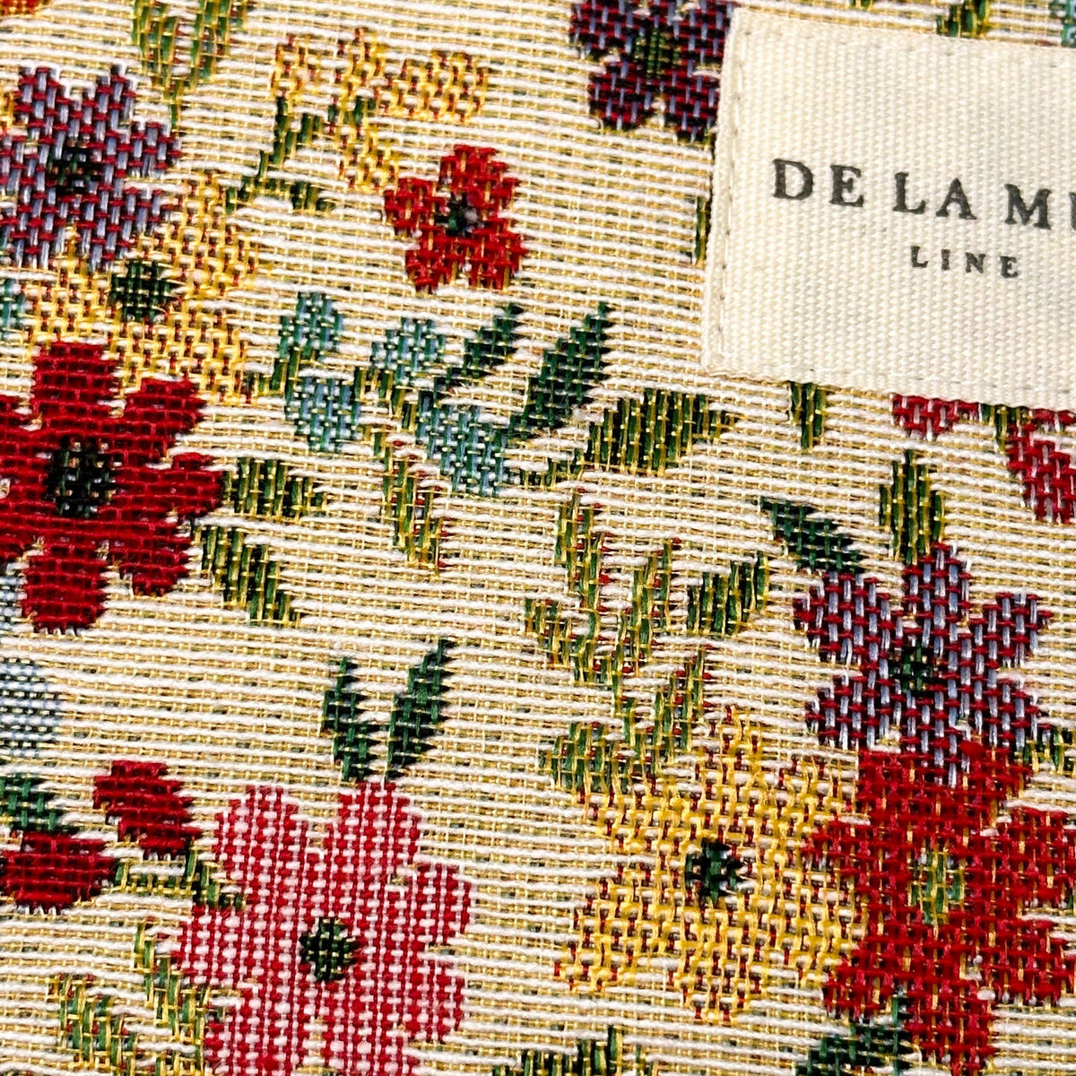 A close-up of the Floral Jacquard Small Makeup Bag / Coin Purse – Beige (Blum) by DE LA MUR LINE US Inc features a colorful woven fabric with a red, yellow, and green floral pattern. In the top right corner, a white label displays the DE LA MUR LINE branding. This stylish textile makes it perfect for use as a coin purse or small makeup bag.