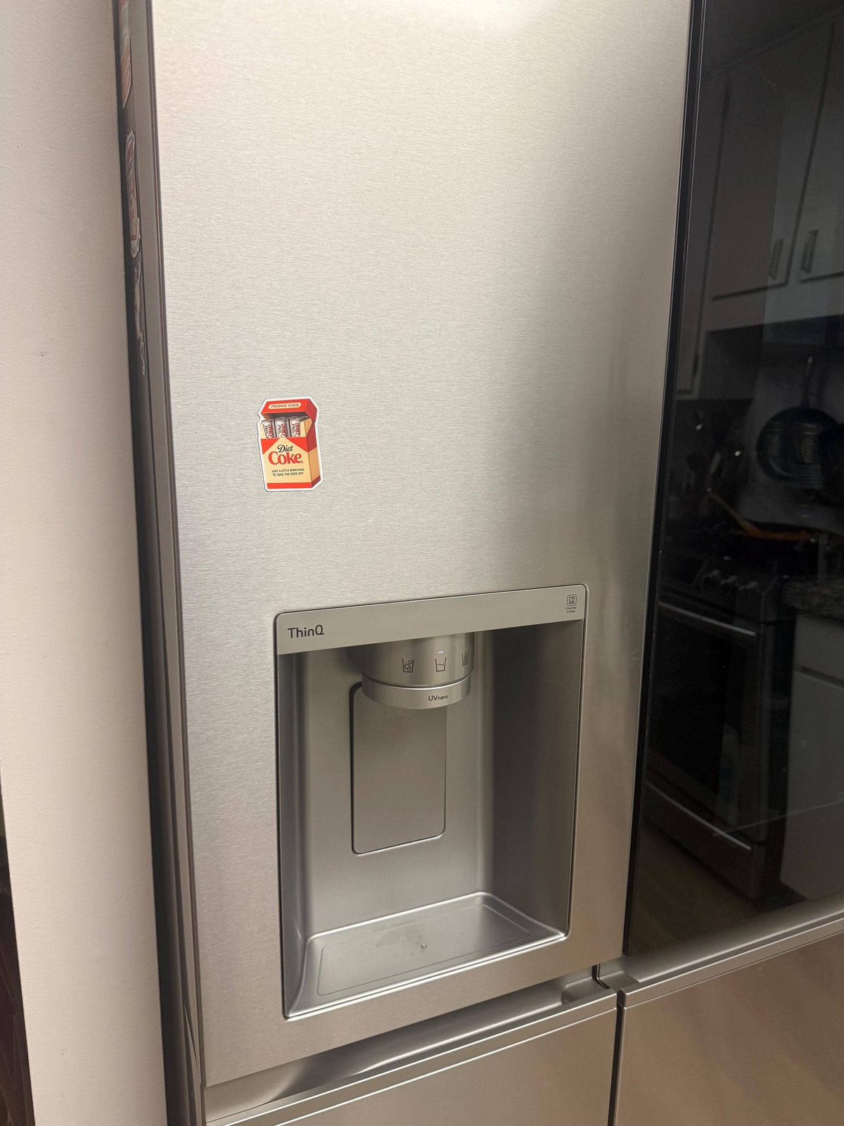 A stainless steel refrigerator with a water and ice dispenser features the Diet Coke Fridge Cigs Sticker by Hello Brand | Sticker My Art on the upper left door. Kitchen cabinets and a stove are visible in the background.