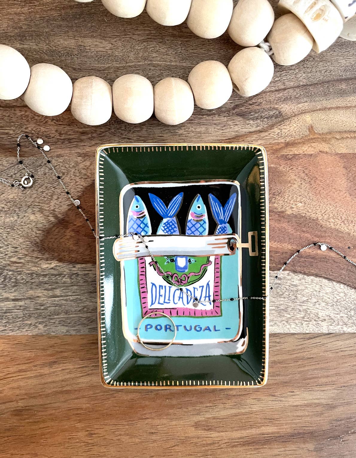 Rectangular dark green Porcelain Trinket Dish - Sardines with gold detailing, featuring a sardine tin design labeled DELICADEZA and PORTUGAL, with a thin silver necklace draped across it.