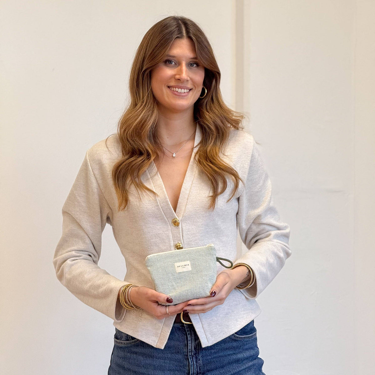 A woman with long wavy hair, dressed in a light beige jacket and blue jeans, smiles as she holds the Striped Small Makeup Bag – Light Green & Green (Teo) by DE LA MUR LINE US Inc, featuring a label and crafted in Barcelona, against a plain off-white background.