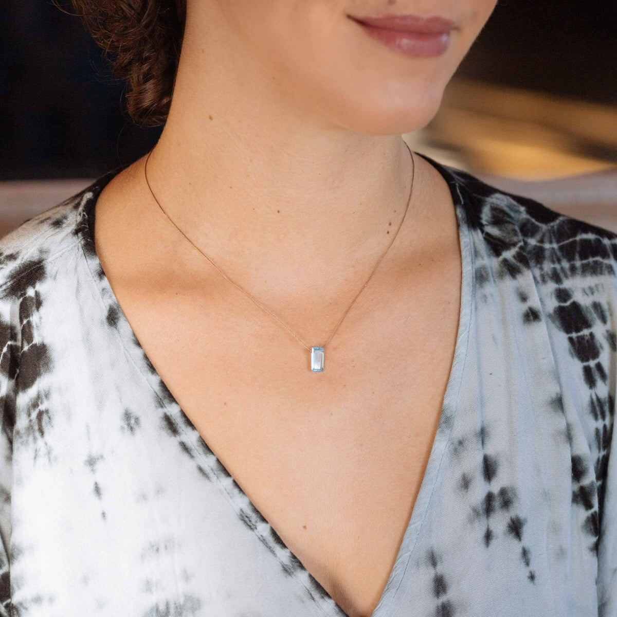 A woman wears a blue and white V-neck top paired with the SoulKu Sky Blue Topaz Refined Necklace for New Beginnings, featuring a delicate handmade rectangular pendant.