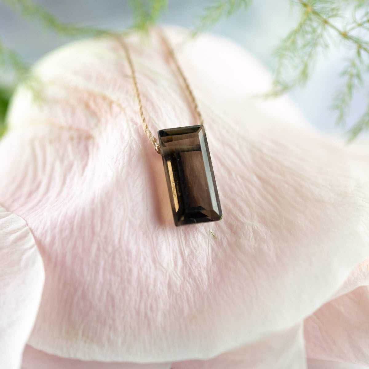The Smoky Quartz Refined Necklace for Release by SoulKu, featuring a genuine rectangular gemstone, sits gracefully on a pale pink flower petal with blurred green foliage in the background.