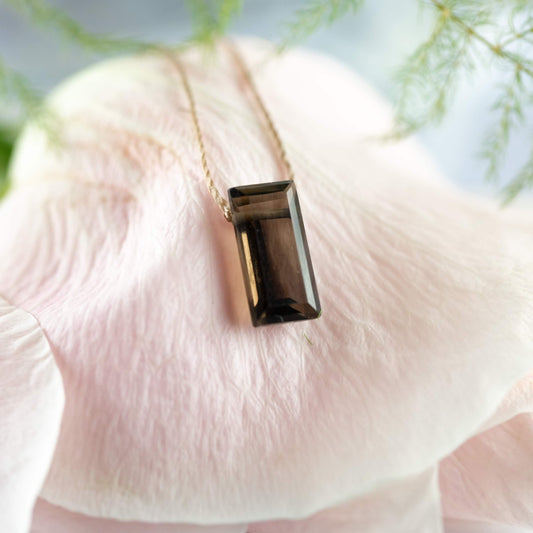 The Smoky Quartz Refined Necklace for Release by SoulKu, featuring a genuine rectangular gemstone, sits gracefully on a pale pink flower petal with blurred green foliage in the background.