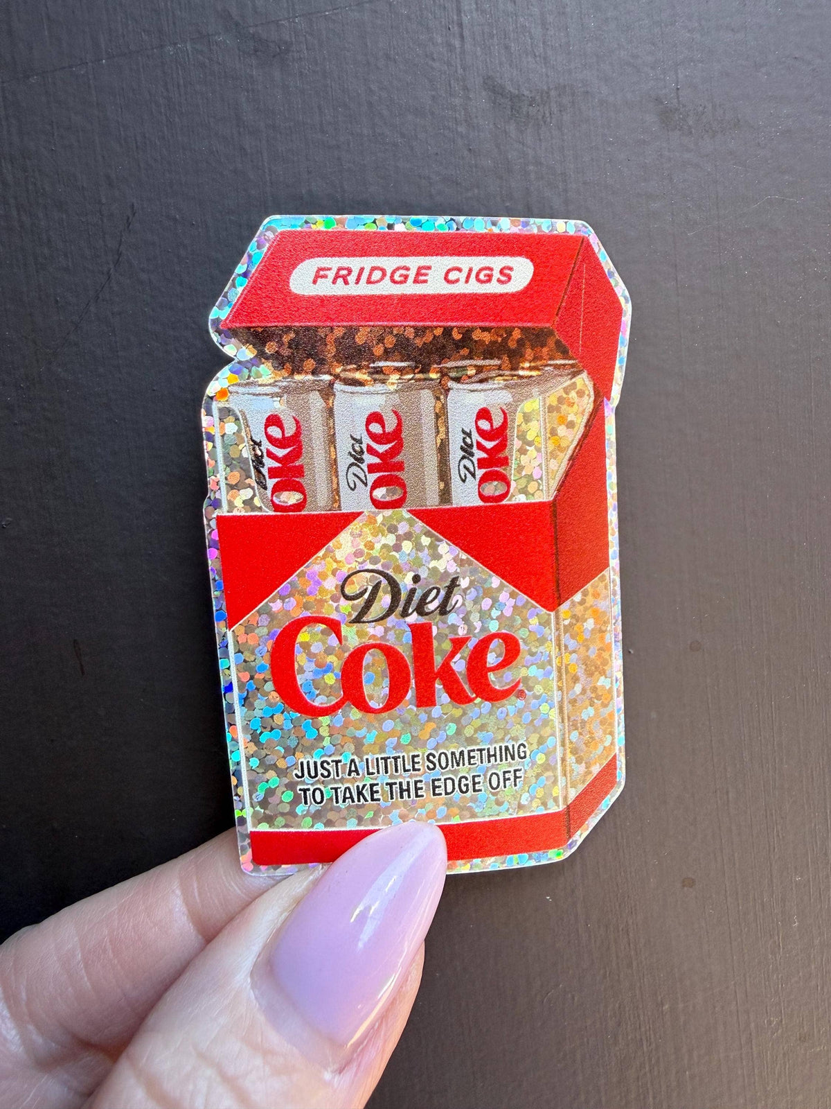 A hand holds the Diet Coke Fridge Cigs Sticker by Hello Brand | Sticker My Art— a sparkly, weather-resistant vinyl sticker showing a Fridge Cigs carton filled with Diet Coke cans and the text: Just a little something to take the edge off.