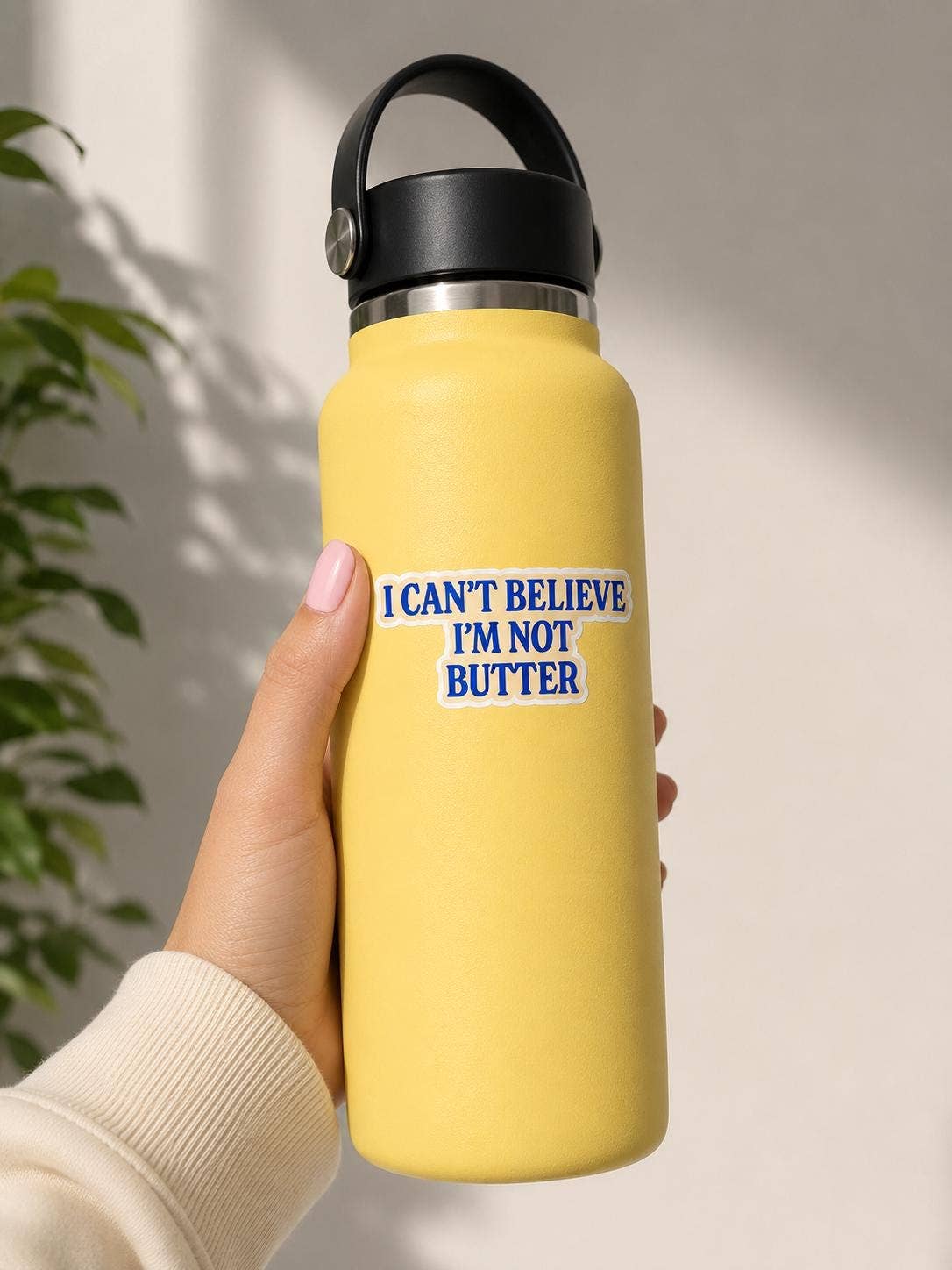 A hand holds a yellow water bottle with an I can’t believe I’m not butter Sticker from Hello Brand | Sticker My Art, made of weather-resistant vinyl and a matte finish. A green plant and soft shadows appear in the background.