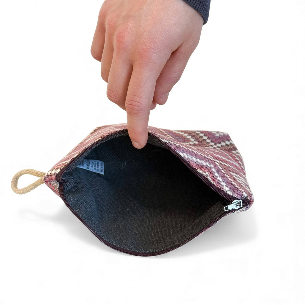 A hand holds open the Geometric Jacquard Small Makeup Bag – Pink & Lilac (Nana) by DE LA MUR LINE US Inc, revealing its dark interior. The exterior features a stylish geometric kilim pattern, and a rope loop on one side makes it perfect for use as a chic makeup bag.