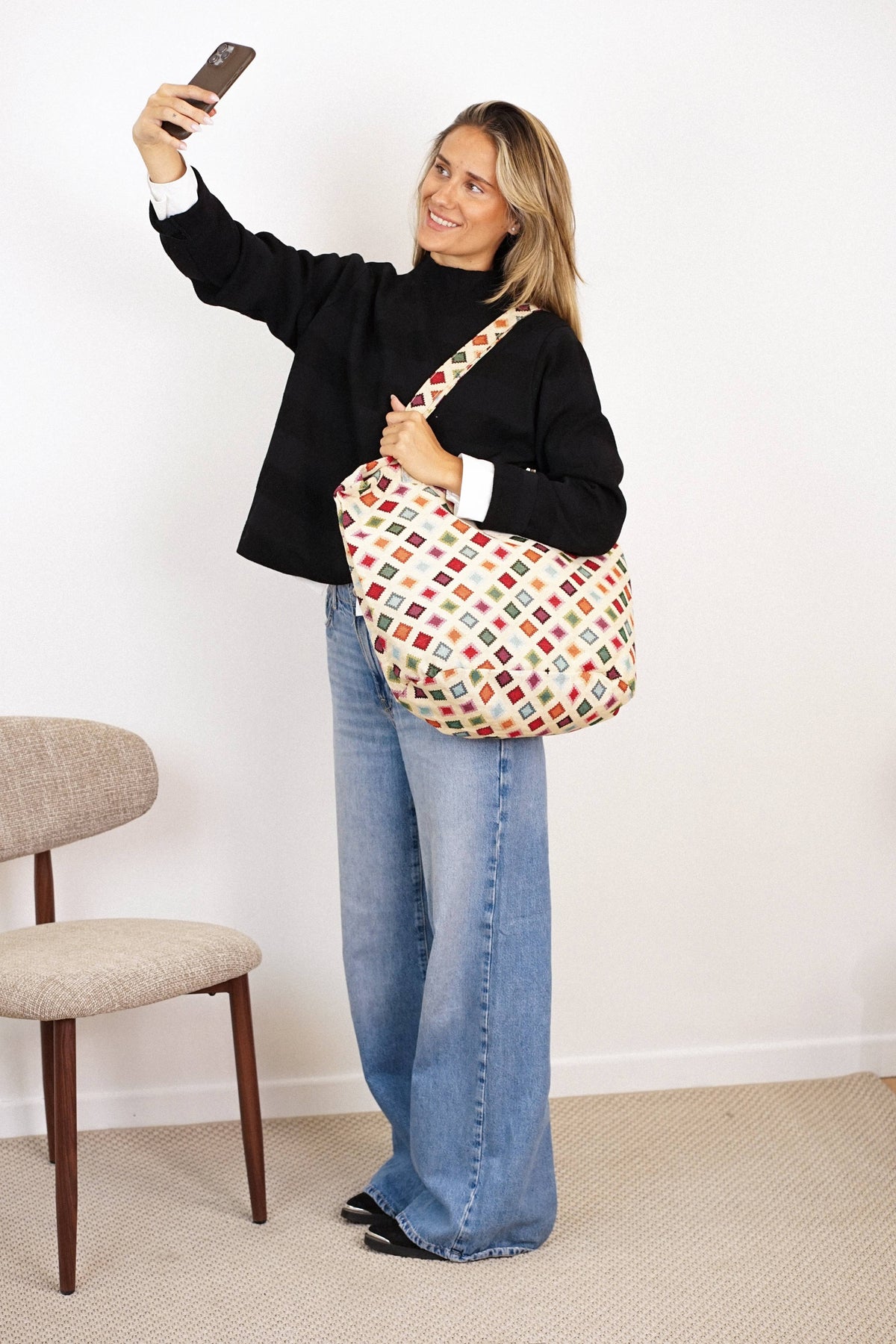 A woman in a black sweater and wide-leg jeans takes a selfie while carrying the Geometric Jacquard Large Zippered Tote Bag – Multicolor (Zac) by DE LA MUR LINE US Inc. She stands next to a beige chair in a minimalist room with white walls and a carpeted floor.