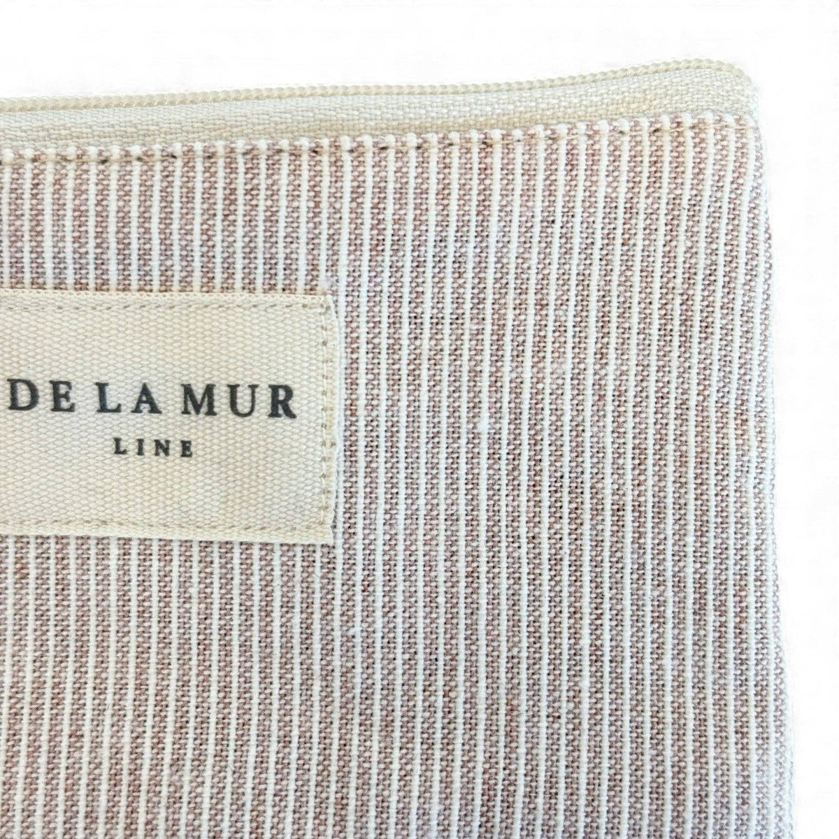 A close-up of the Striped Coin Purse – Gray & Cream, featuring beige and white striped jacquard fabric and a rectangular label with “DE LA MUR LINE US Inc” in black text, highlighting the elegant craftsmanship made in Barcelona.