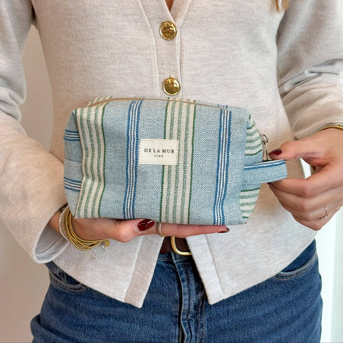 A person in a light cardigan and jeans holds the Striped Jacquard Squared Makeup Bag – Blue & Green (Cata) from DE LA MUR LINE US Inc, made from luxurious jacquard fabric and featuring blue and green stripes. Gold bracelets adorn their wrist, and their nails are painted dark red.