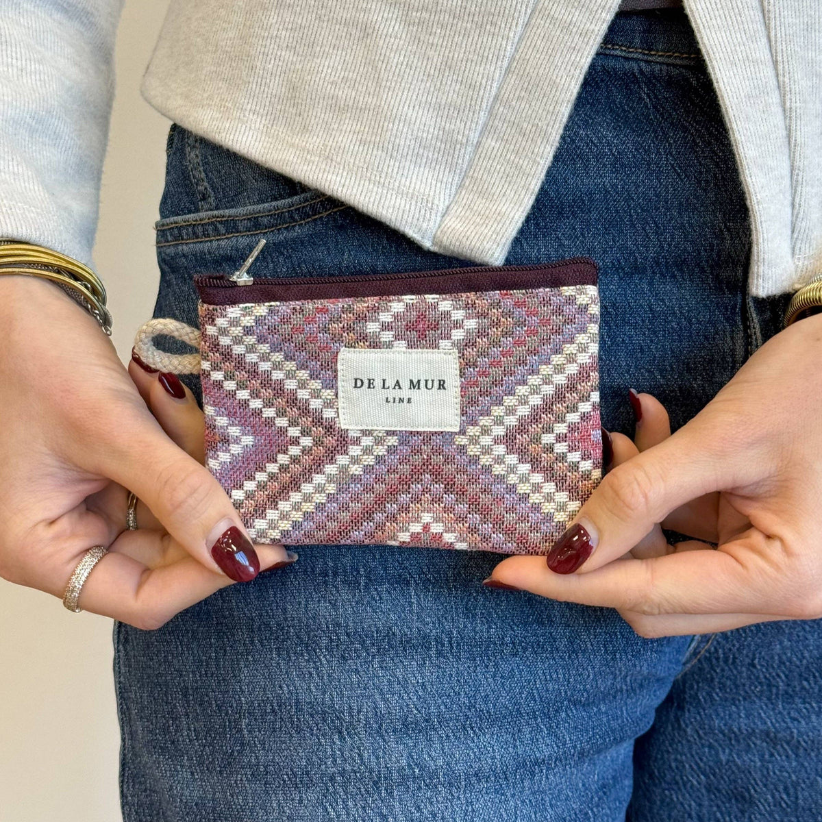 A person in jeans and a light top holds the Geometric Jacquard Coin Purse – Pink & Lilac (Nana) by DE LA MUR LINE US Inc with both hands.