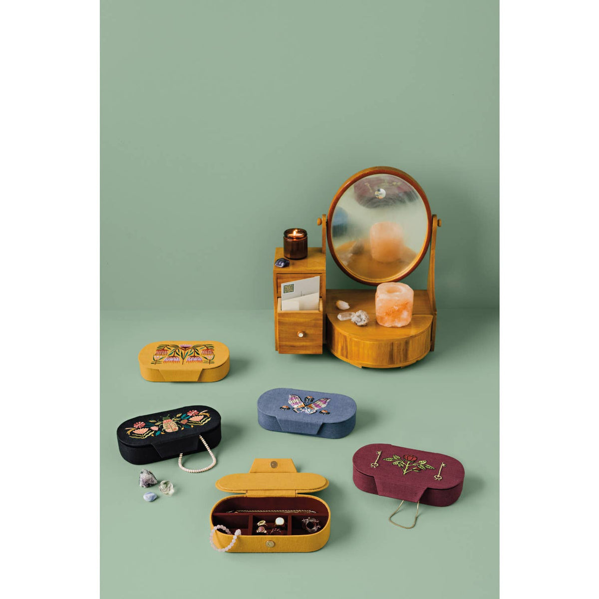 Five small, embroidered Amulet Embroidered Jewelry Box - Black/Pink amulets in yellow, black, blue, and dark red arranged on a surface below a wooden vanity mirror.