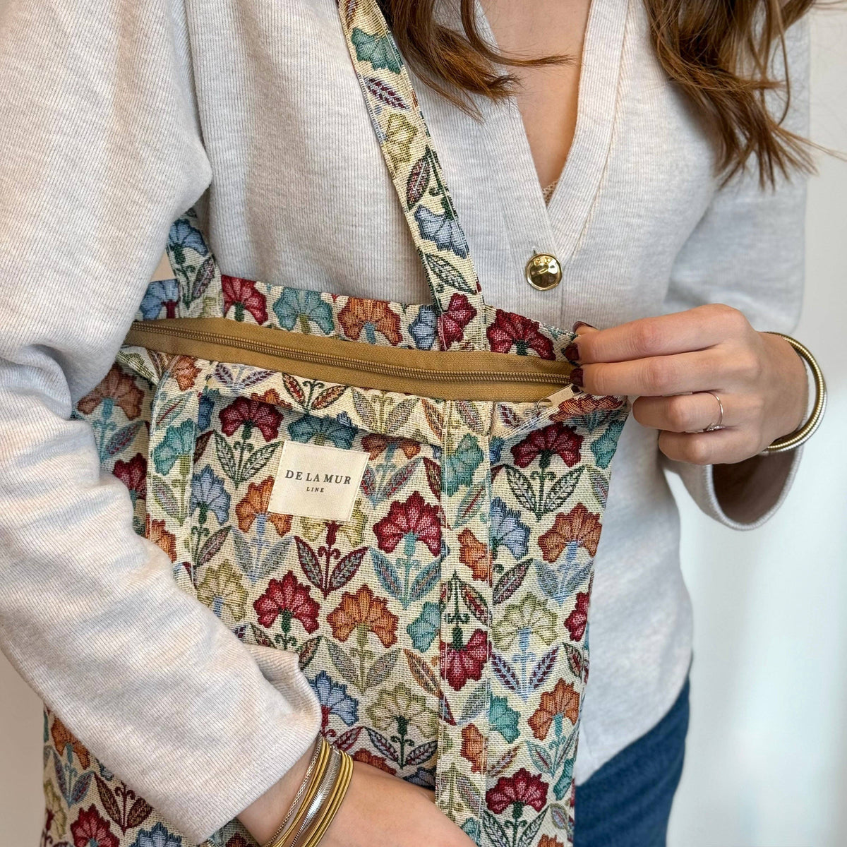 A woman in a light sweater holds the Floral Jacquard Medium Zippered Tote Bag – Multicolor (Flora) by DE LA MUR LINE US Inc, unzipping the main compartment. She accessorizes with gold jewelry on her wrist and fingers.