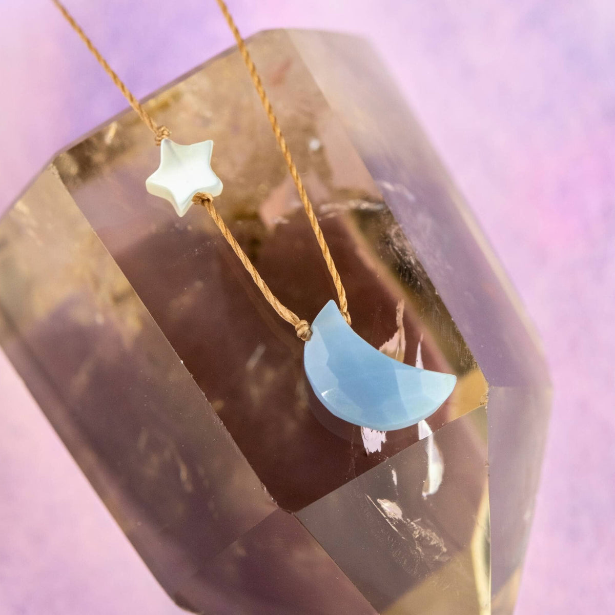 A SoulKu Blue Opal - I Love You To The Moon & Back - Necklace featuring a light blue opal crescent moon pendant and a small white star bead on a thin brown cord, resting on a large clear crystal.