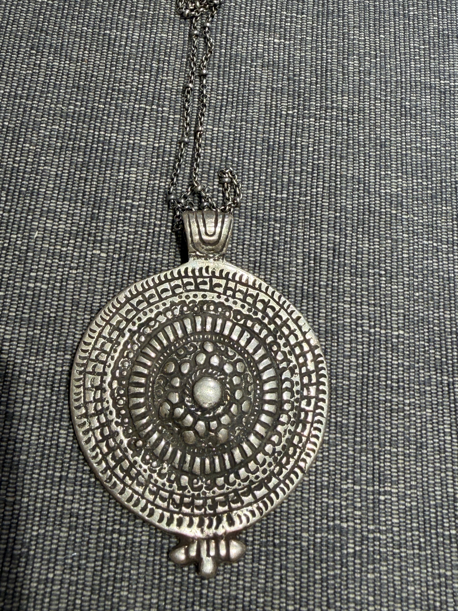 The Large Geo Silver Pendant Necklace by Mosaic Sun Designs features a bold, round silver pendant with intricate geometric patterns, suspended from a matching silver chain and showcased against a gray textured fabric background.