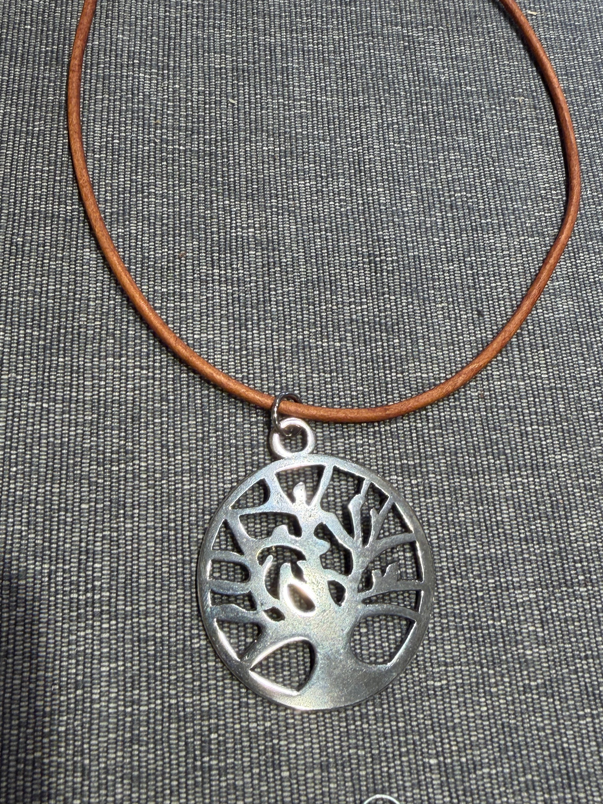 The Leather Tree of Life Lg Pendant by Mosaic Sun Designs features a round silver pendant with a tree motif, suspended from a brown leather cord necklace and displayed on textured gray fabric.