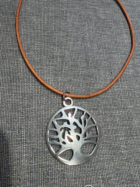The Leather Tree of Life Lg Pendant by Mosaic Sun Designs features a round silver pendant with a tree motif, suspended from a brown leather cord necklace and displayed on textured gray fabric.
