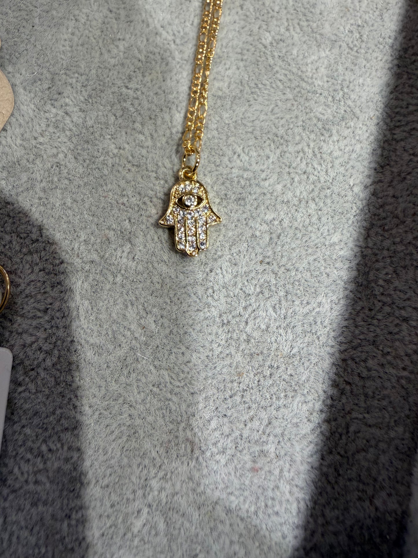 The DIYH Hamsa CZ Necklace - GF features a gold chain and a Hamsa hand pendant with clear stones, shown on a light gray textured surface.