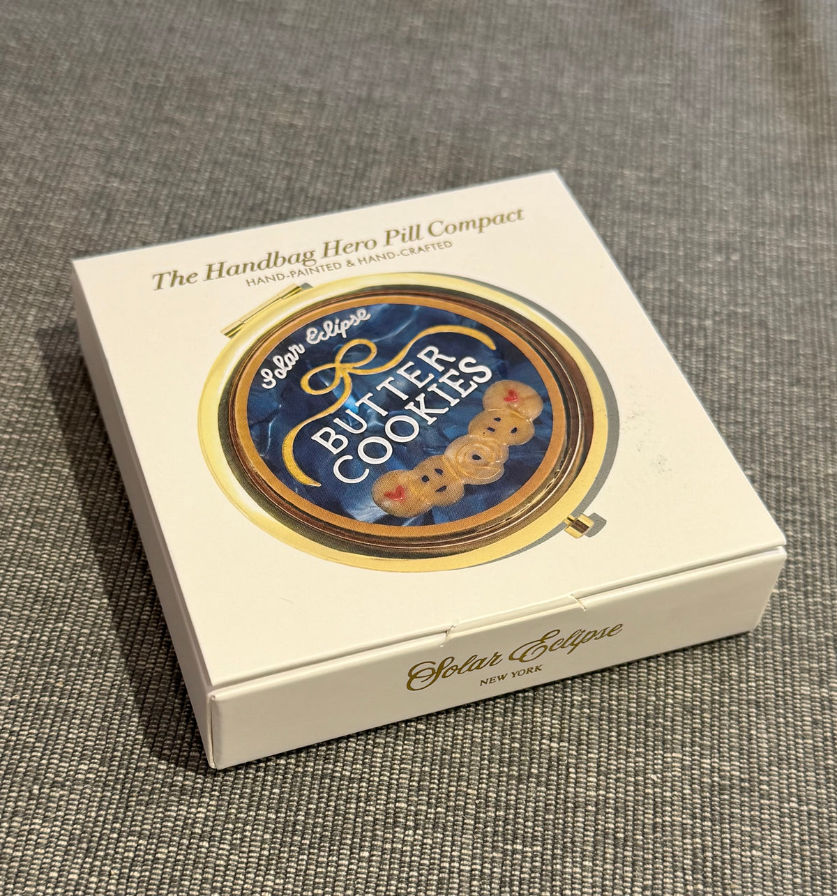 A white box containing a round, gold-rimmed Butter Cookies Handbag Hero Pill Compact - Hand-Painted with a label that features a blue background, a gold ribbon, and the text Butter Cookies above an illustration of three small cookies.