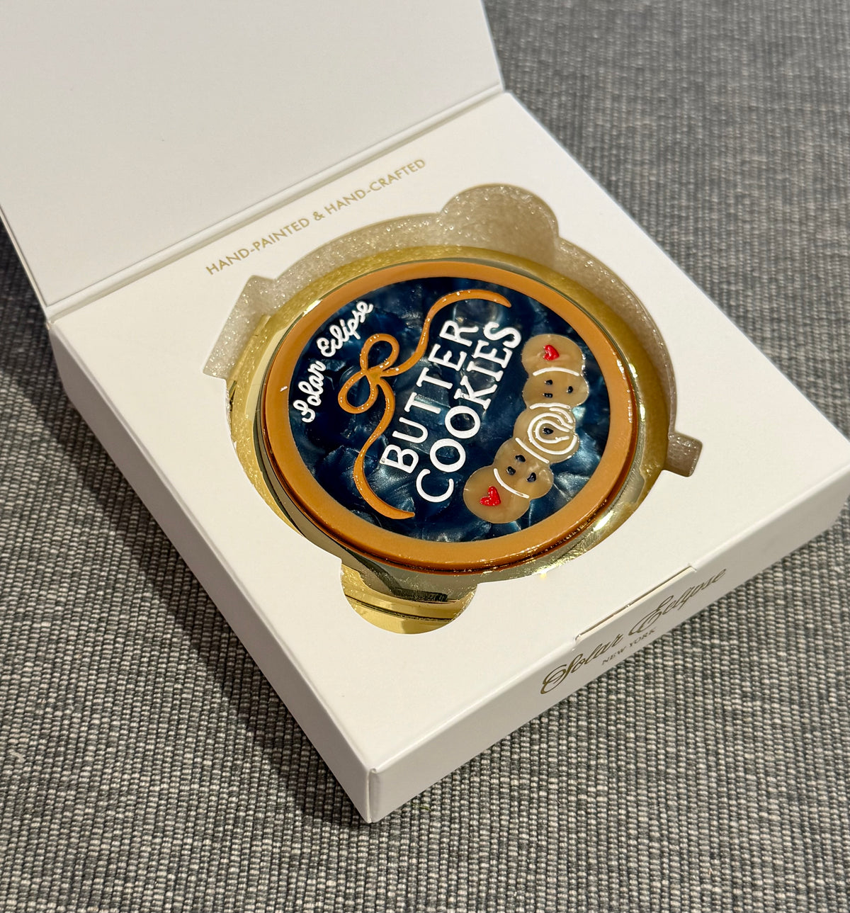 A round, vintage-inspired Butter Cookies Handbag Hero Pill Compact - Hand-Painted in an open white box, featuring a dark blue lid decorated with hand-painted butter cookies and the text Butter Cookies.