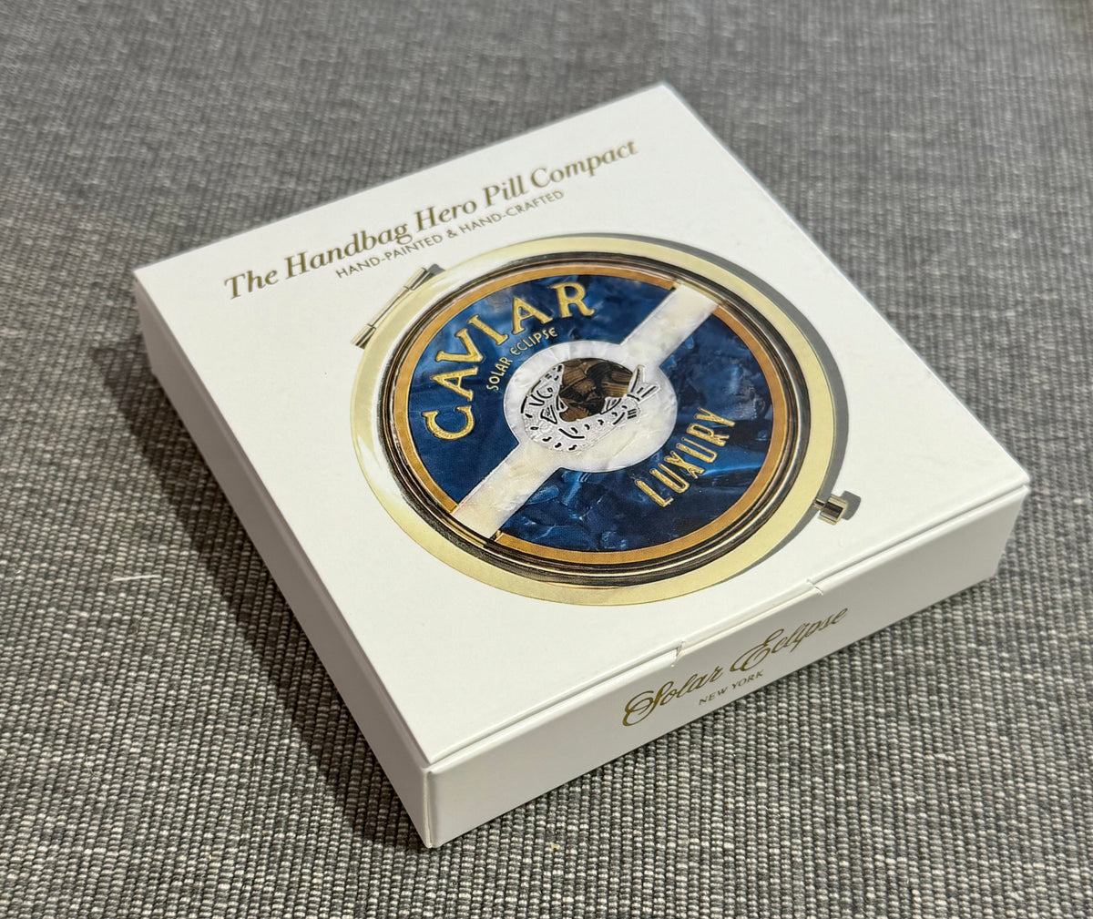 A round, hand-painted Solar Eclipse Caviar Handbag Hero Pill Compact featuring a blue and white design with the text CAVIAR and LUXURY, resting inside its branded white box.