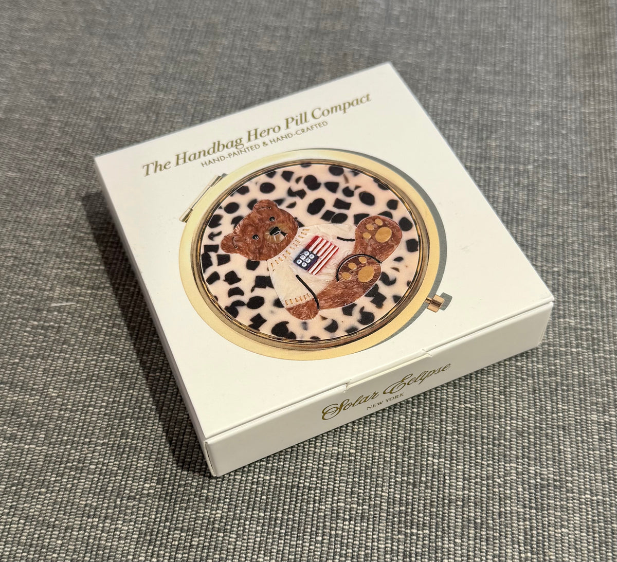 A Solar Eclipse Teddy Bear Americana Handbag Hero Pill Compact featuring a hand-painted teddy bear wearing an American flag sweater on a leopard print background in a white vintage-inspired case.