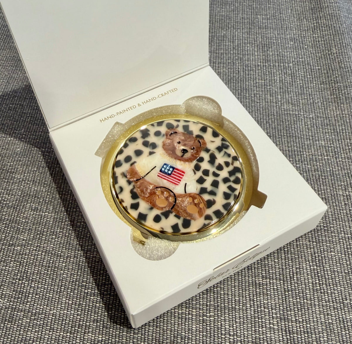 Solar Eclipse Teddy Bear Americana Handbag Hero Pill Compact - Hand-Painted, a round compact pill case featuring a hand-painted teddy bear wearing a sweater with an American flag, housed in an open white gift box.