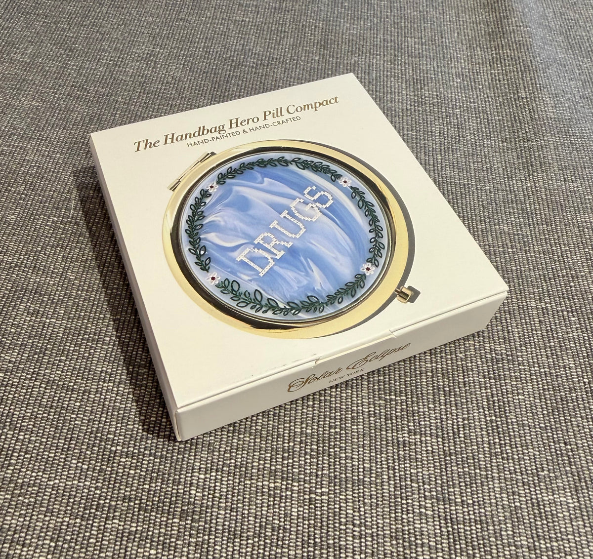 A Solar Eclipse Cross-Stitch Drugs Handbag Hero Pill Compact - Hand - Painted featuring a round gold case with a blue, cloud-patterned lid stitched with the word DRUGS in white, displayed in its product box.
