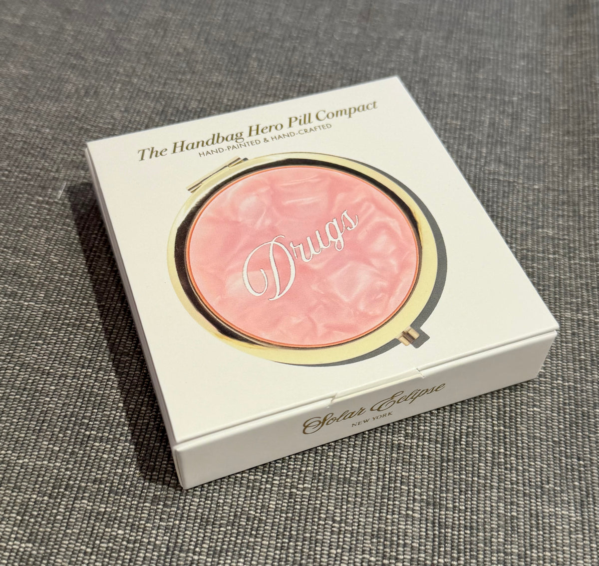 White square product box featuring a gold-rimmed Solar Eclipse Pink Drugs Handbag Hero Pill Compact - Hand - Painted with a swirling pink surface labeled Drugs in white cursive script.
