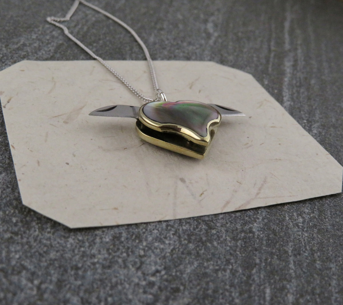 Mini Heart Knife Necklace featuring a heart-shaped locket with abalone shell inlay on a sterling silver curb chain, with two small folding knife blades extended from the sides.