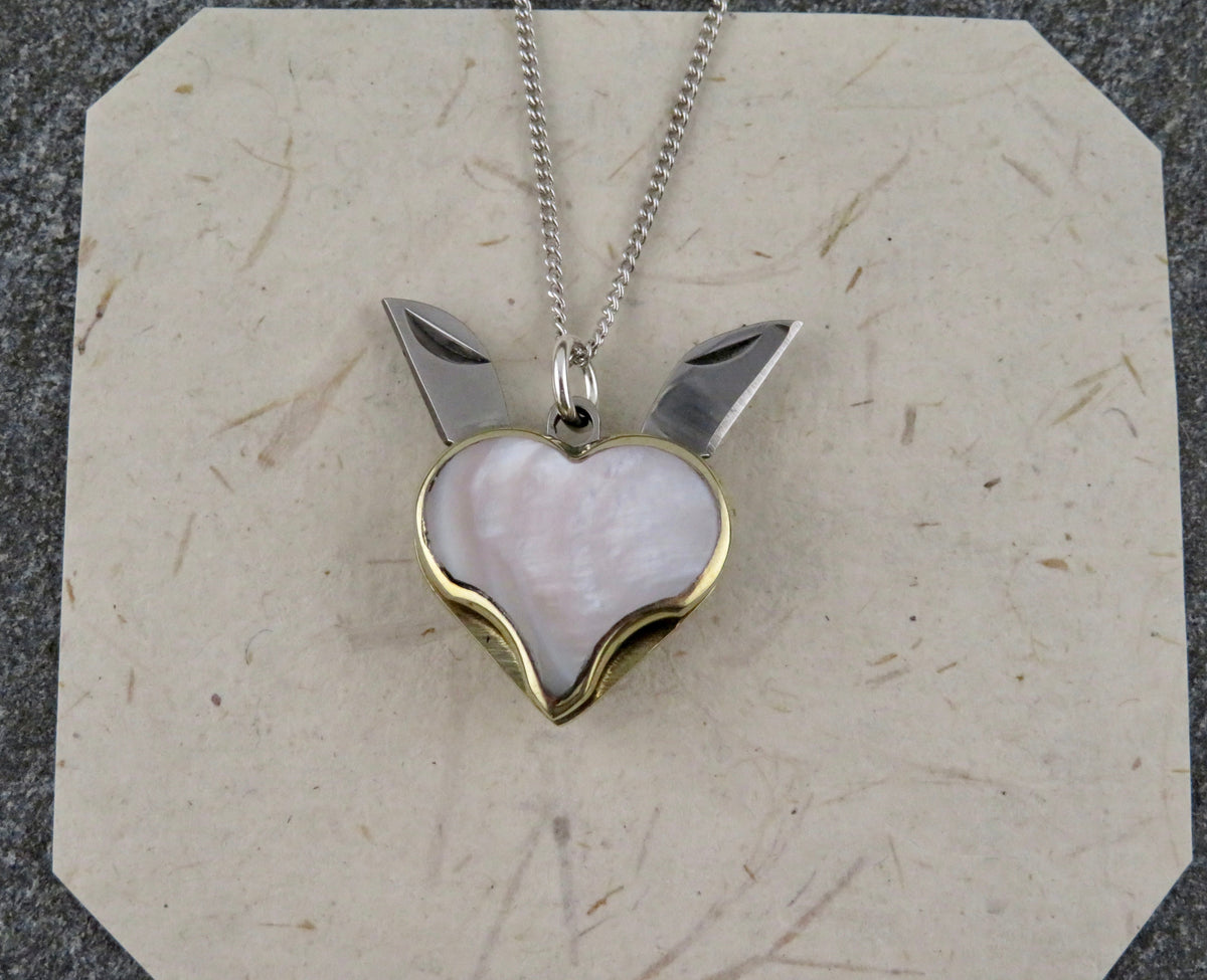 A Blue Ridge Knives Mini Heart Knife Necklace featuring a heart-shaped pendant with an abalone shell inlay and two small folding blades extending from the top, hanging on a sterling silver curb chain.