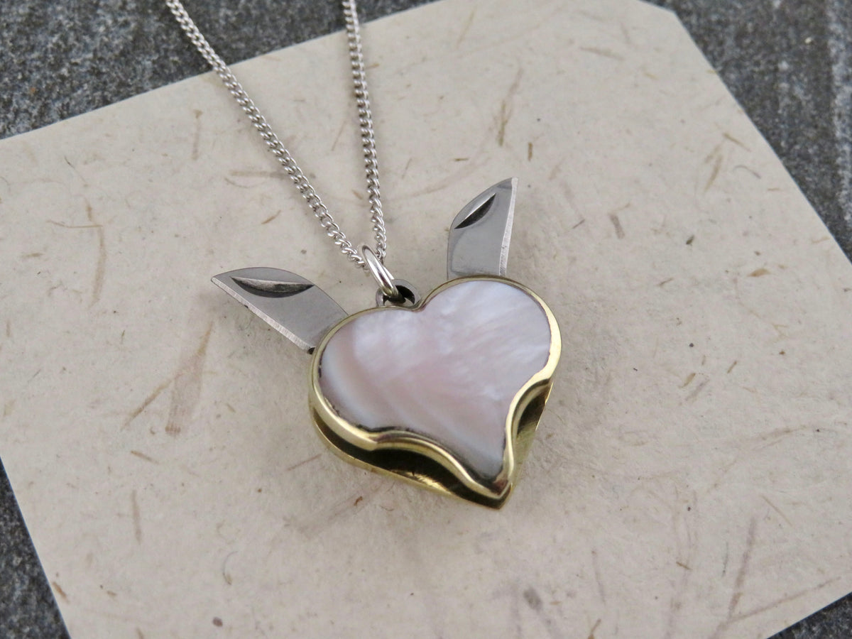 A Blue Ridge Knives Mini Heart Knife Necklace featuring a heart-shaped pendant with an abalone shell inlay and two small folding knife blades, hanging from a sterling silver curb chain.