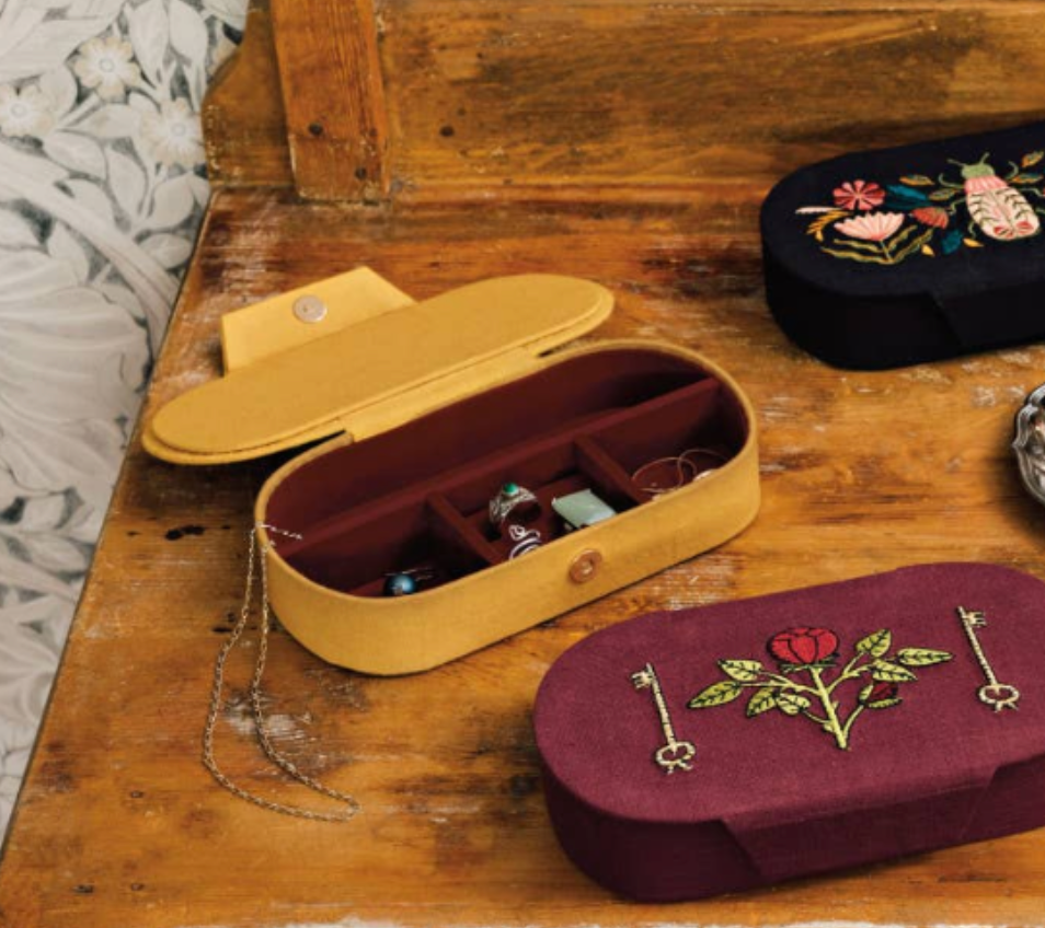 An open yellow rectangular Venus Embroidered Jewelry Box - Yellow/Burgundy with a burgundy interior sits next to two closed boxes, one burgundy with rose embroidery and one dark blue with insect and floral embroidery.