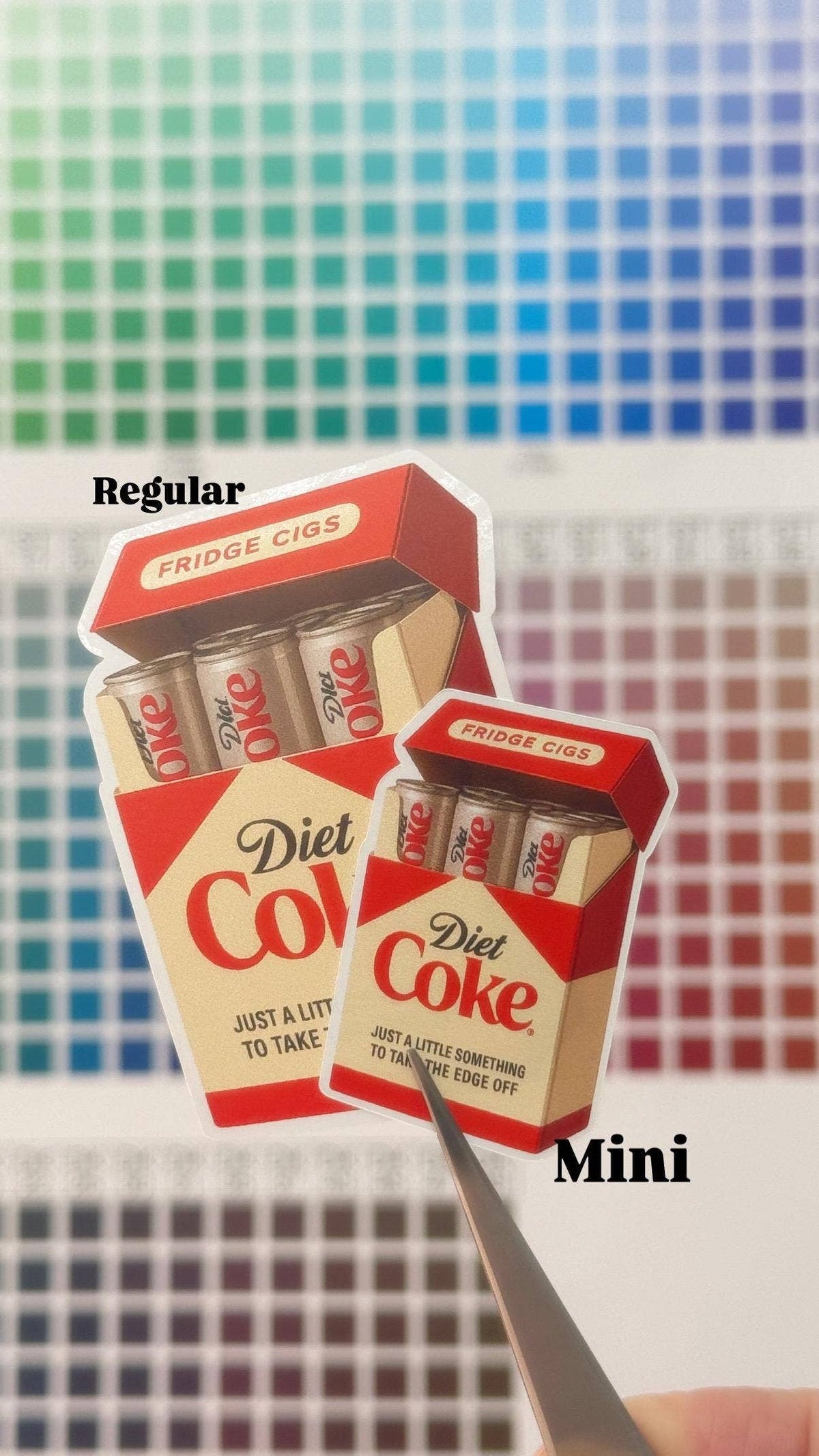 A hand holds a mini weather-resistant Diet Coke Fridge Cigs Sticker by Hello Brand | Sticker My Art next to a regular-sized one, both displayed on a colorful square-patterned background.