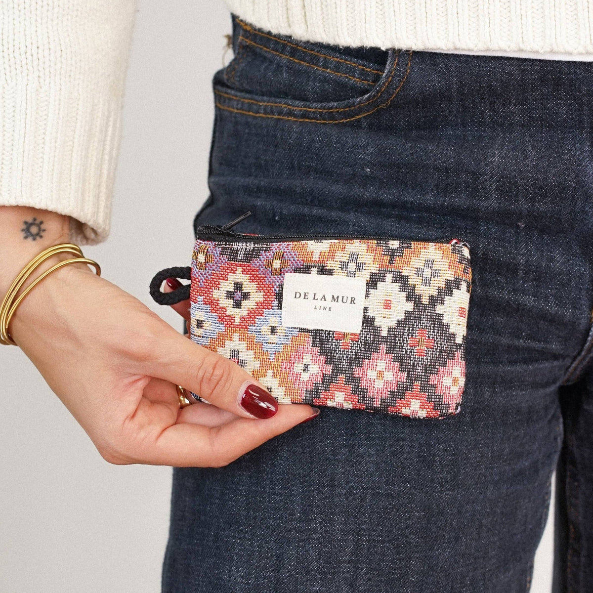 A person dressed in dark jeans and a white sweater holds the Geometric Jacquard Coin Purse – Multicolor (Ore) by DE LA MUR LINE US Inc. They accessorize with gold bangles and have a small star tattoo on their wrist.