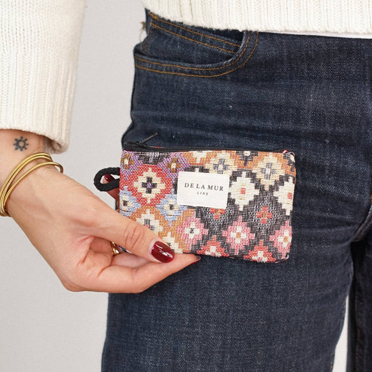 A person dressed in dark jeans and a white sweater holds the Geometric Jacquard Coin Purse – Multicolor (Ore) by DE LA MUR LINE US Inc. They accessorize with gold bangles and have a small star tattoo on their wrist.