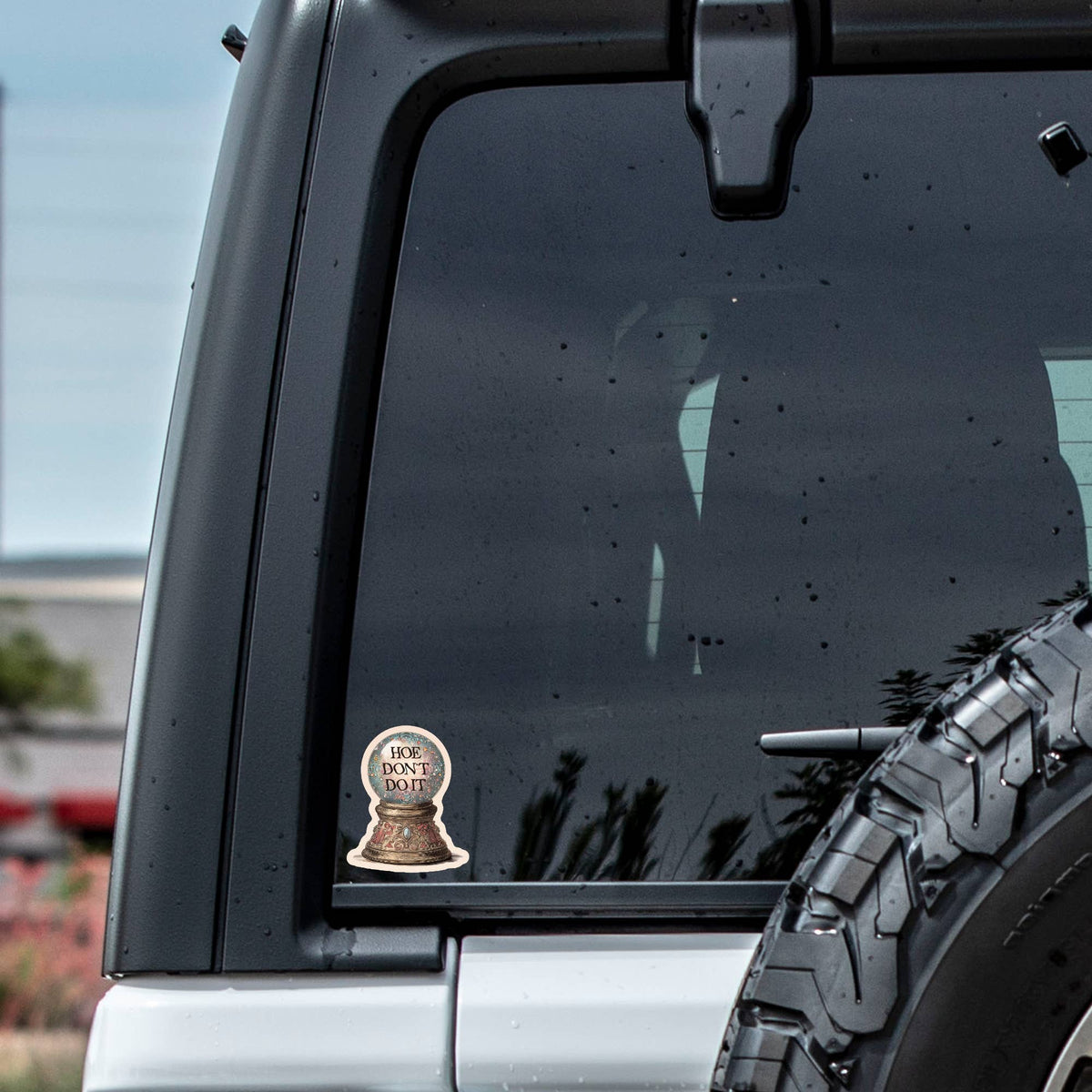 A gray car’s rear window displays the Hoe Don’t Do It Sticker by Juju and Moxie, with raindrops on the glass, a spare tire, and blurred scenery visible in the background.
