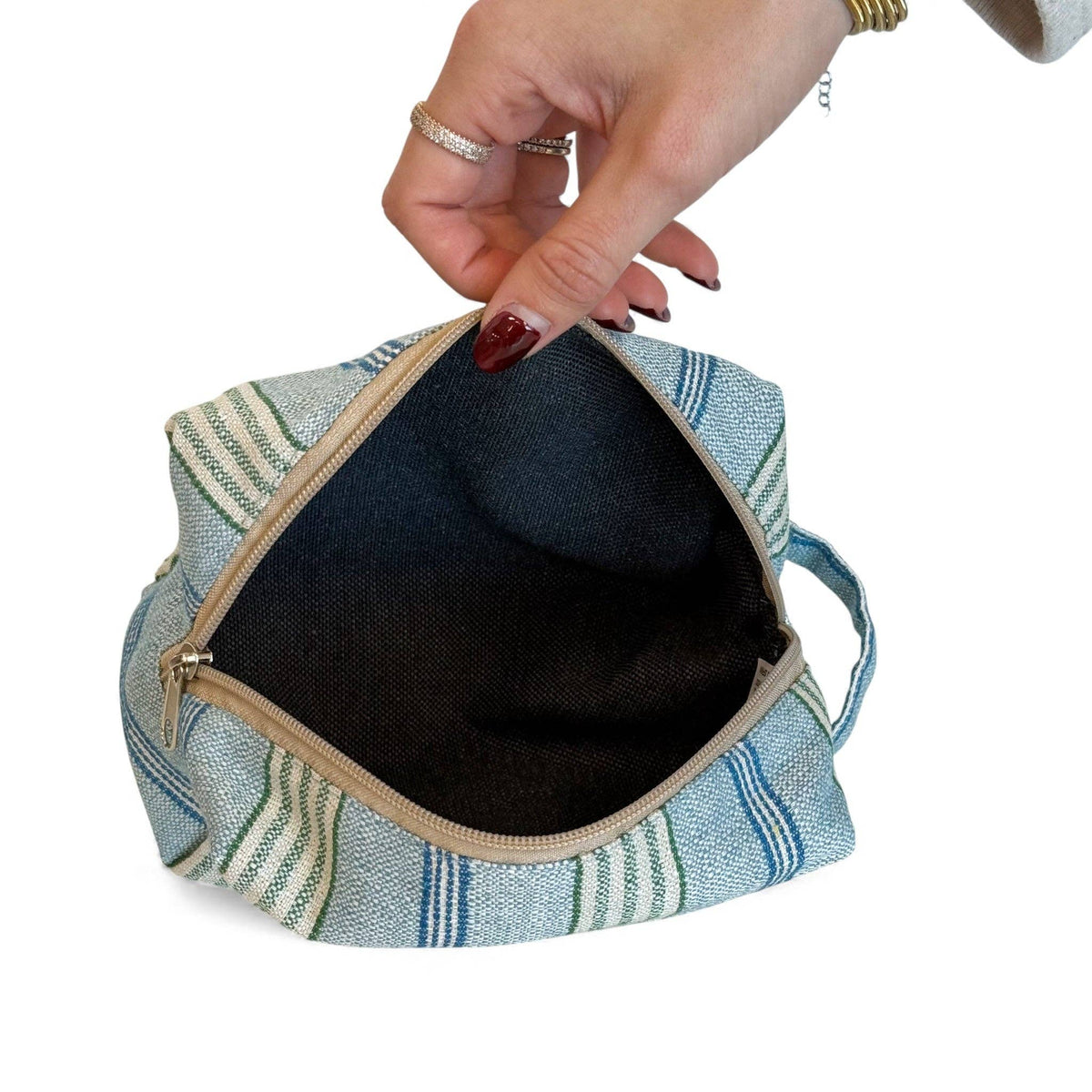 A hand with painted nails holds open the Striped Jacquard Squared Makeup Bag – Blue & Green (Cata) by DE LA MUR LINE US Inc, expertly crafted from blue and green striped jacquard fabric in Barcelona.
