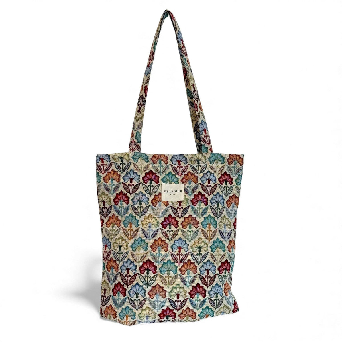 The Floral Jacquard Medium Zippered Tote Bag – Multicolor (Flora) by DE LA MUR LINE US Inc is crafted from jacquard fabric and features two handles, showcasing a vibrant floral leaf design in red, blue, orange, and green tones. A small rectangular label is centered on the plain white background of the bag.