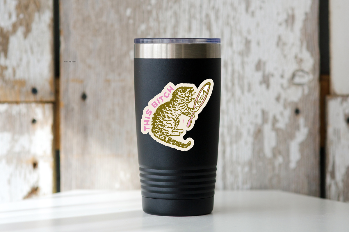 A matte black tumbler on a white surface features the Yarnival This Bitch Cat Sticker, showing a vintage cat holding a mirror with arched pink text, set against rustic weathered wood.