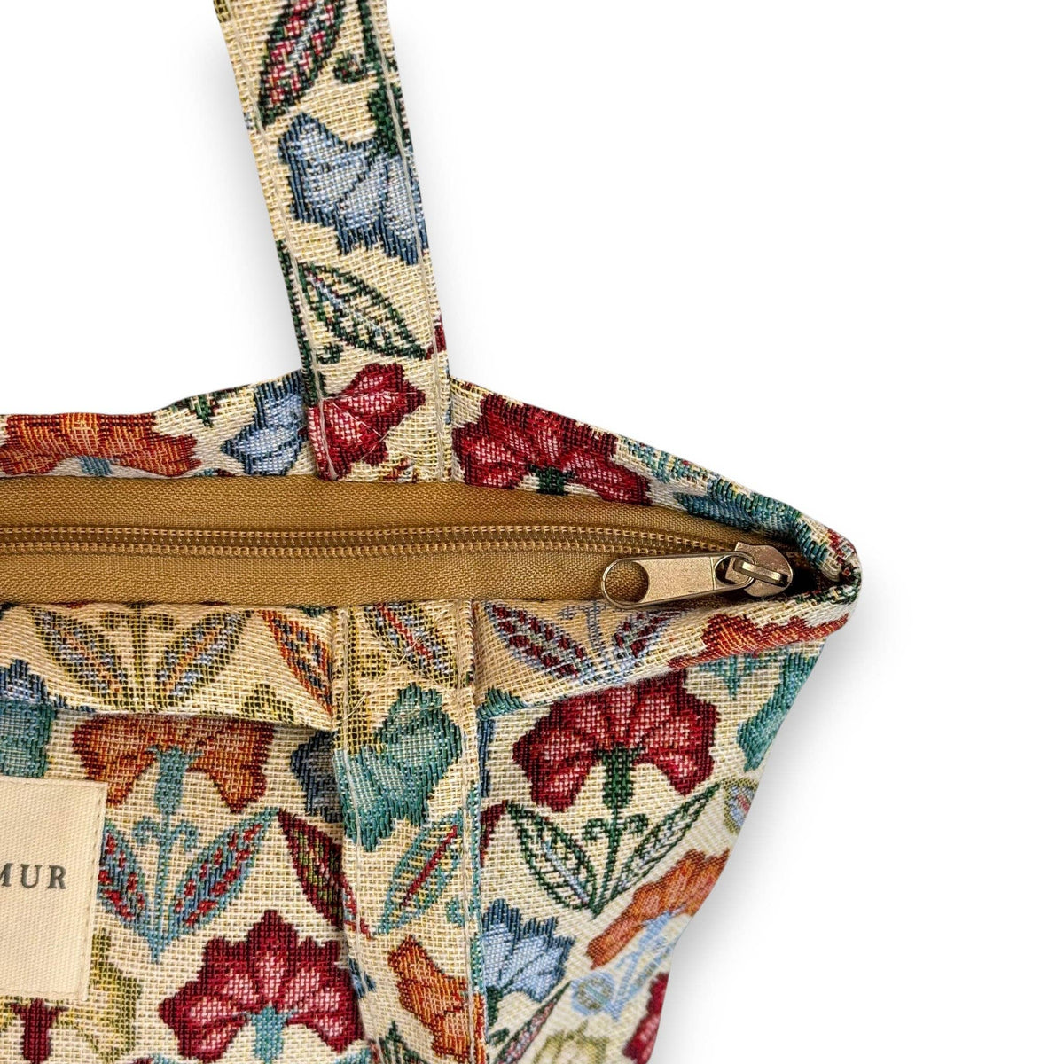 A close-up of the Floral Jacquard Medium Zippered Tote Bag – Multicolor (Flora) by DE LA MUR LINE US Inc showcases its vibrant floral jacquard fabric, complemented by a tan zipper and matching strap. A small partial label appears on the left side, subtly indicating that this beautifully crafted tapestry bag is made in Barcelona.